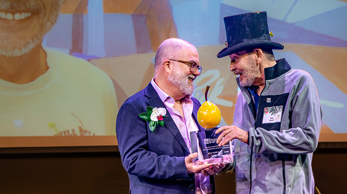 Two men smile together onstage while presenting a Golden Mango Award, with a portrait of the honoree projected behind them.
