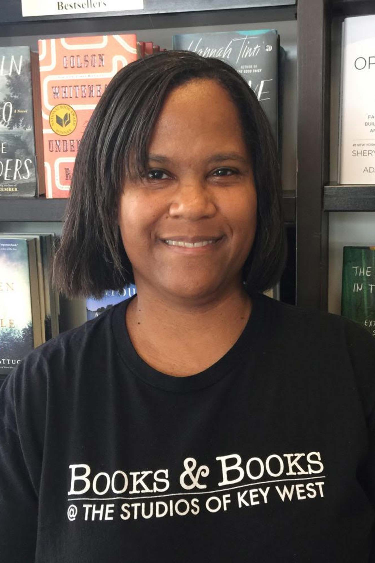 Smiling woman with a chin-length bob wearing a black Books & Books at The Studios of Key West T-shirt, photographed in front of bookstore shelves.