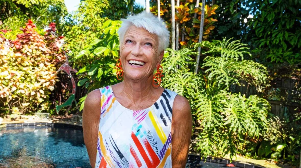 Rosi Ware smiles in a lush tropical garden, wearing a colorful patterned dress and bright orange earrings beside a small pool.
