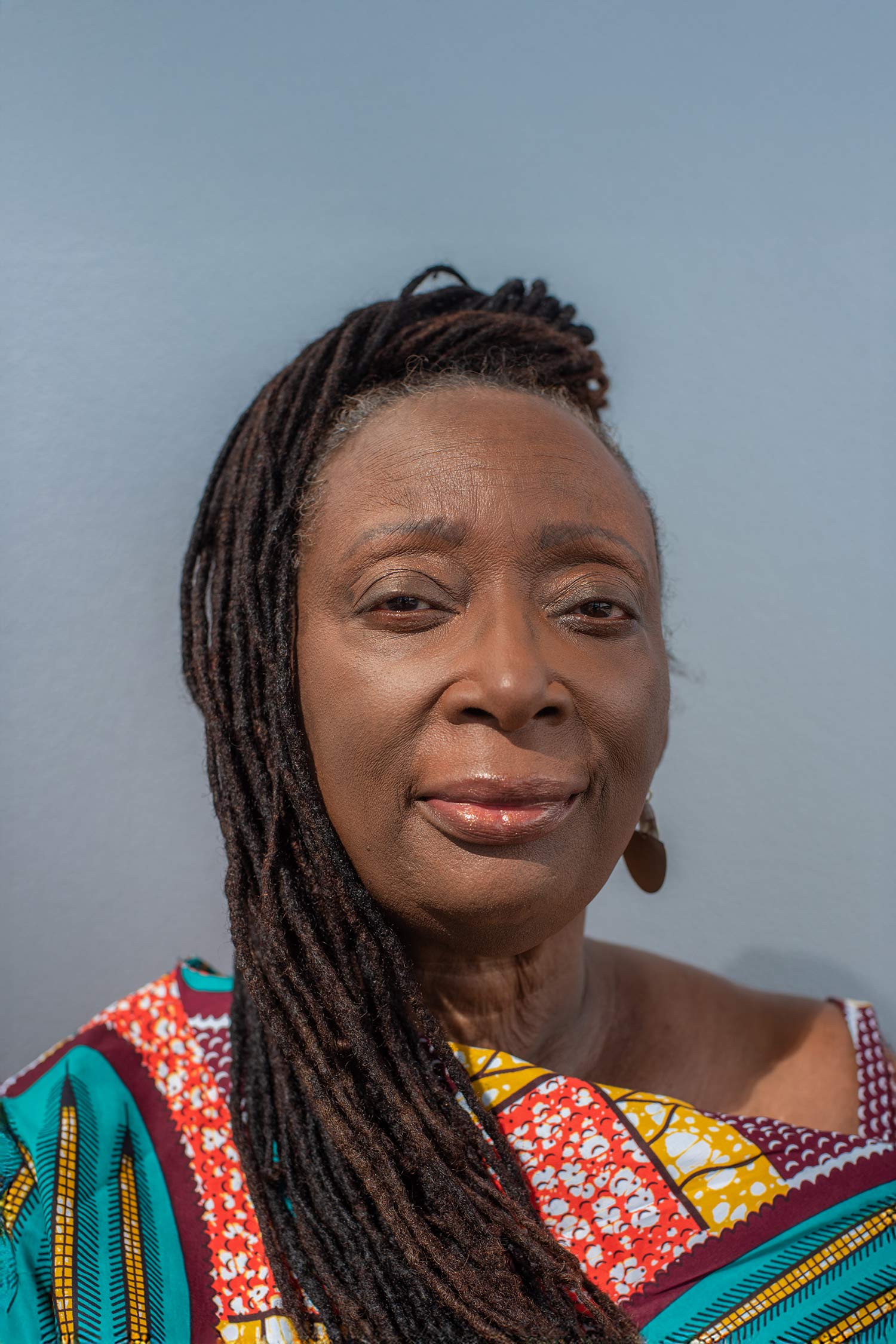 Sandra Jackson-Opoku poses against a blue-gray background wearing a colorful patterned garment, with long locs swept over one shoulder.
