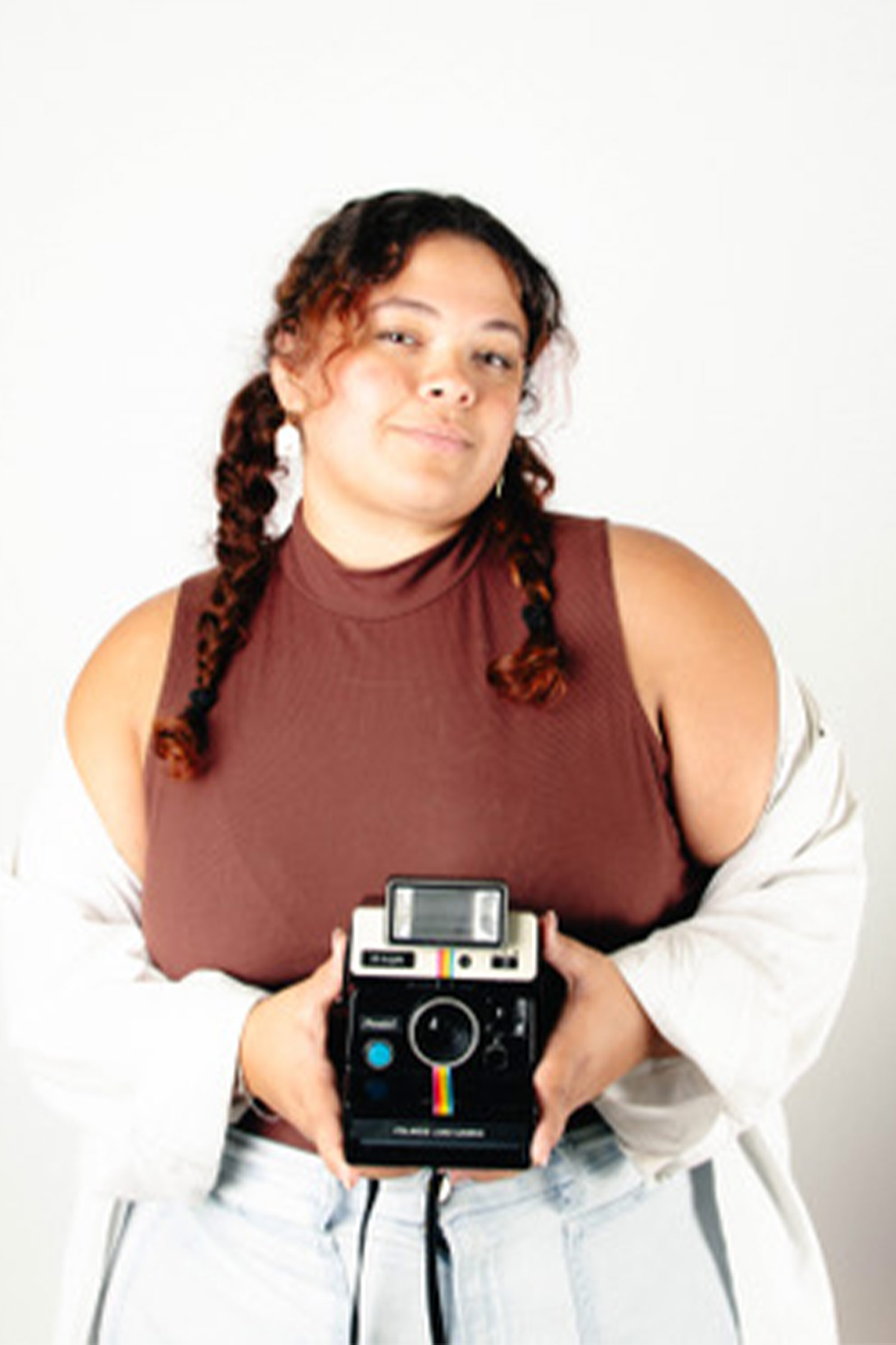Woman with braided hair holds a vintage instant camera while posing against a white background.