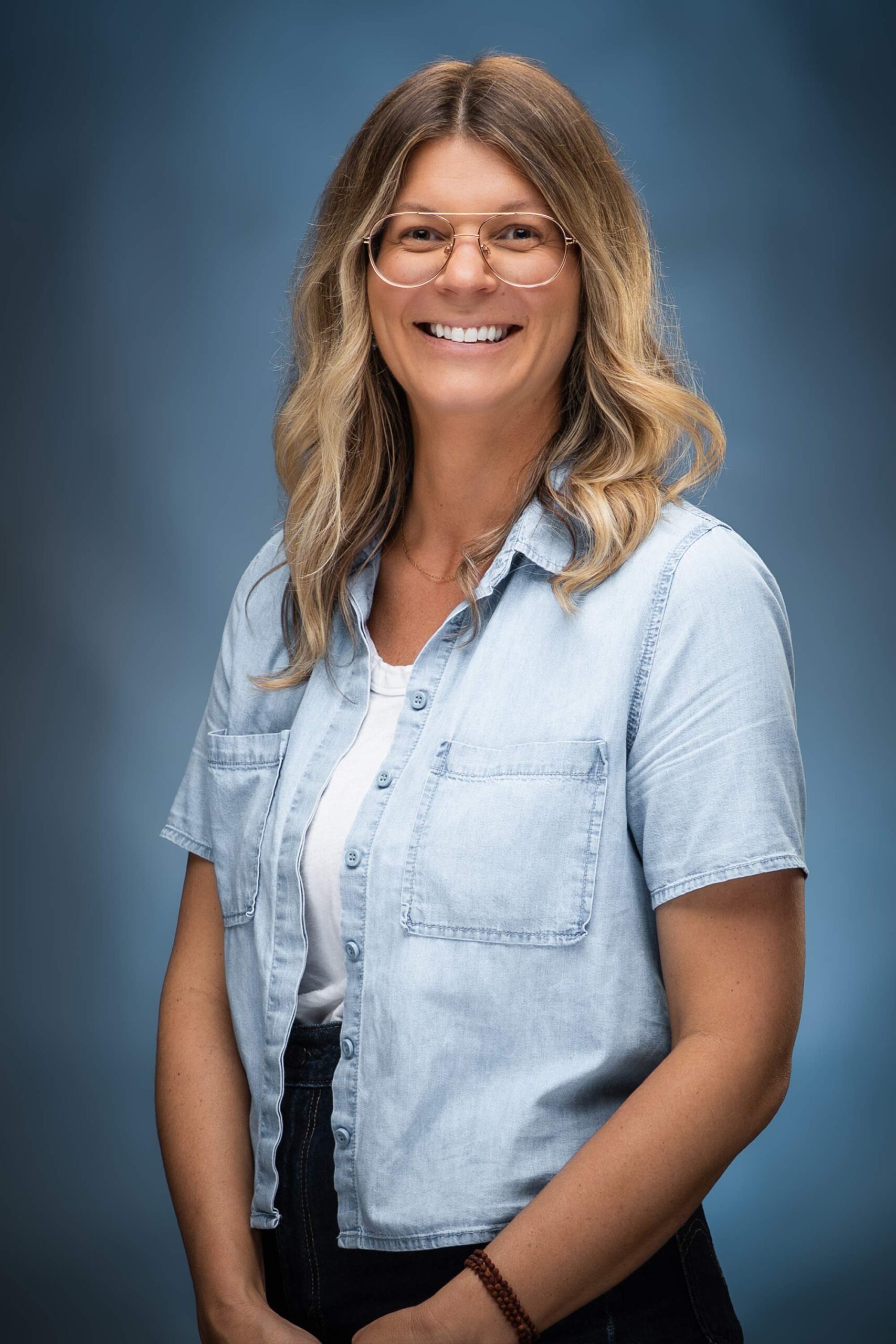 Portrait of Sara smiling in glasses and a light blue button-down shirt against a blue studio background.