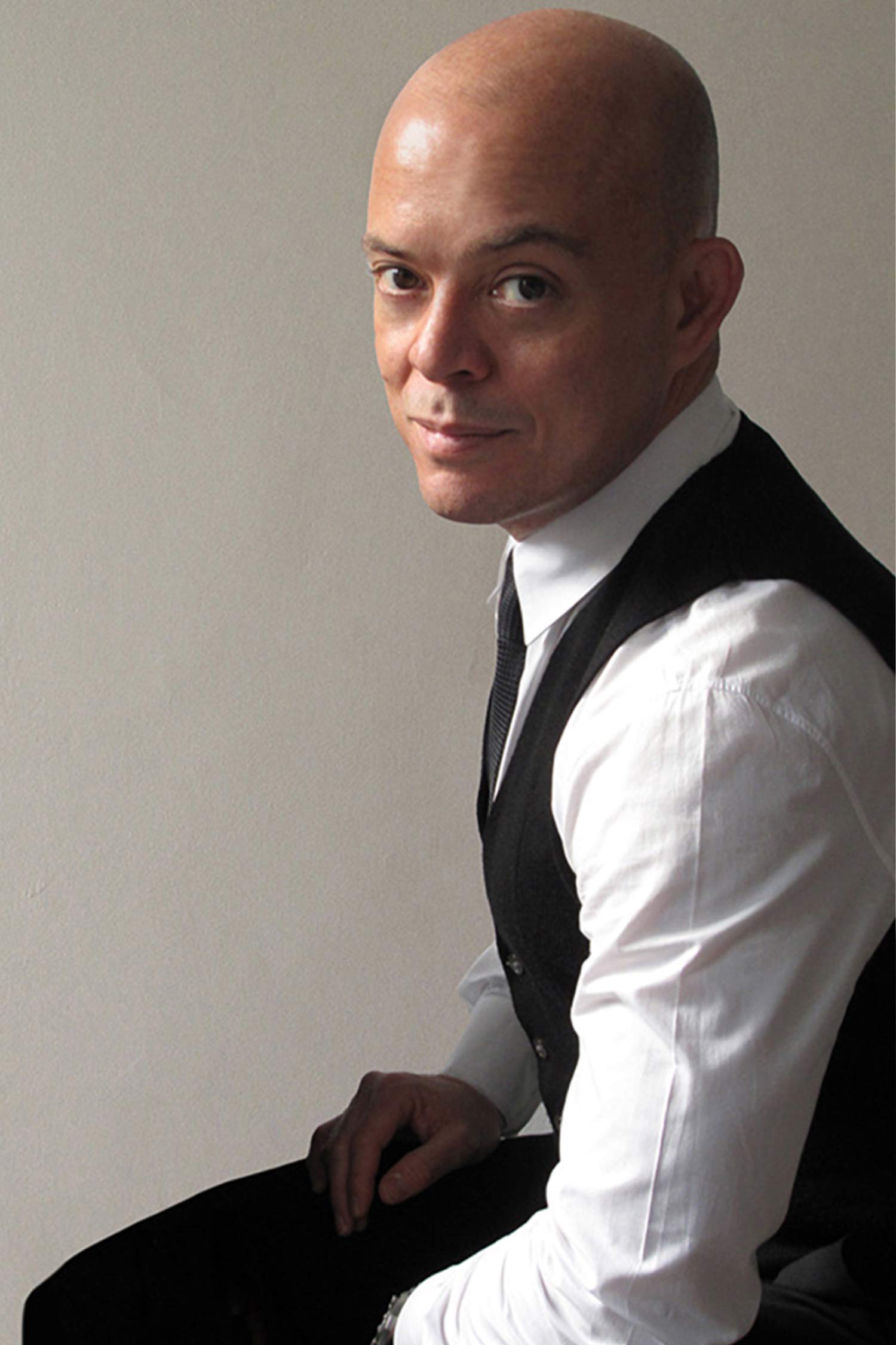Portrait of a seated bald man wearing a white dress shirt, black vest and black tie, turned slightly toward the camera against a plain gray background.