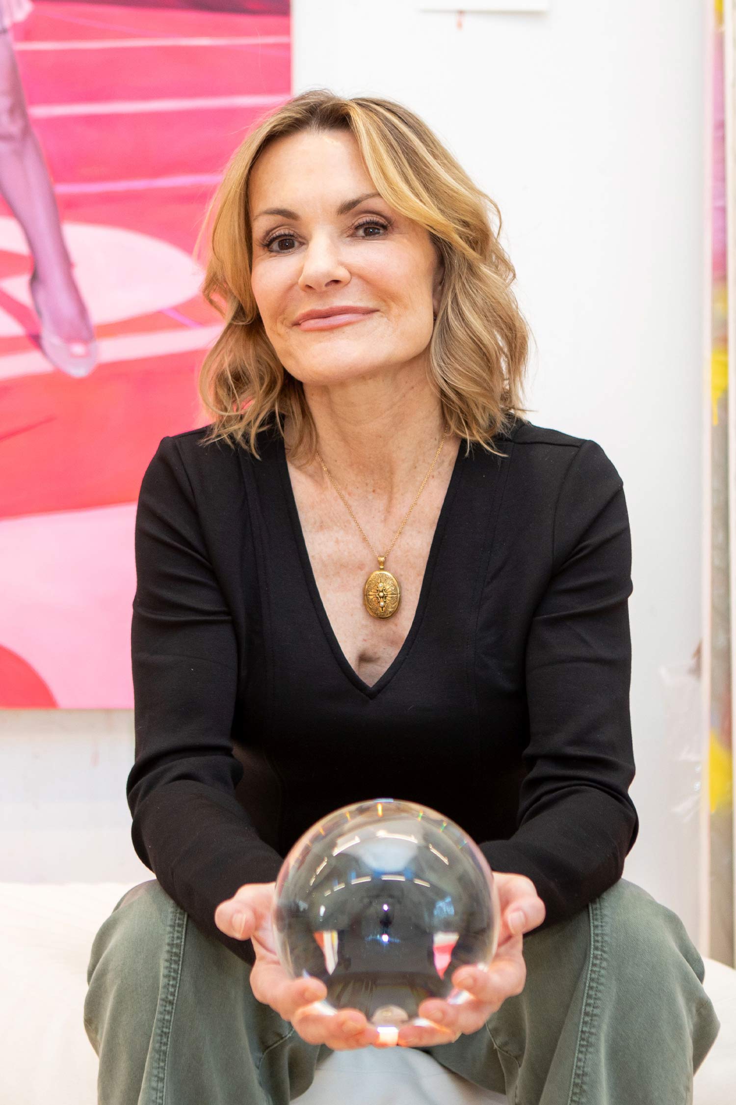 Portrait of a blonde woman seated in an art-filled studio, wearing a black top and holding a large clear glass sphere in both hands.