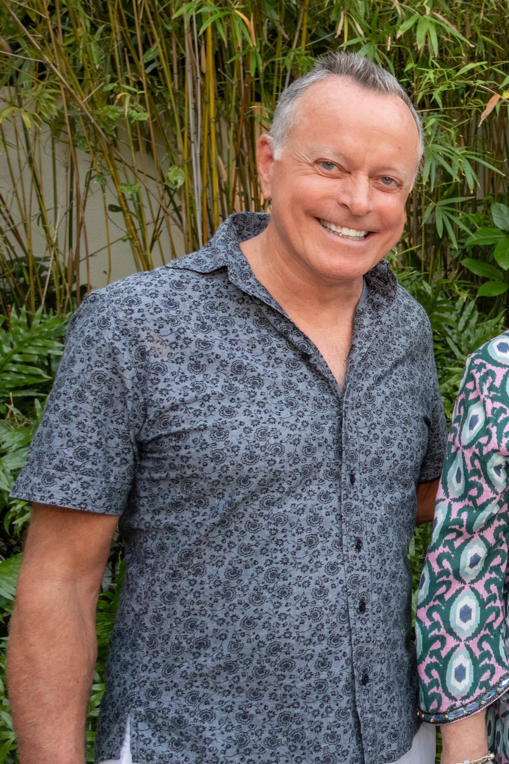 Man smiling outdoors in front of tropical greenery.