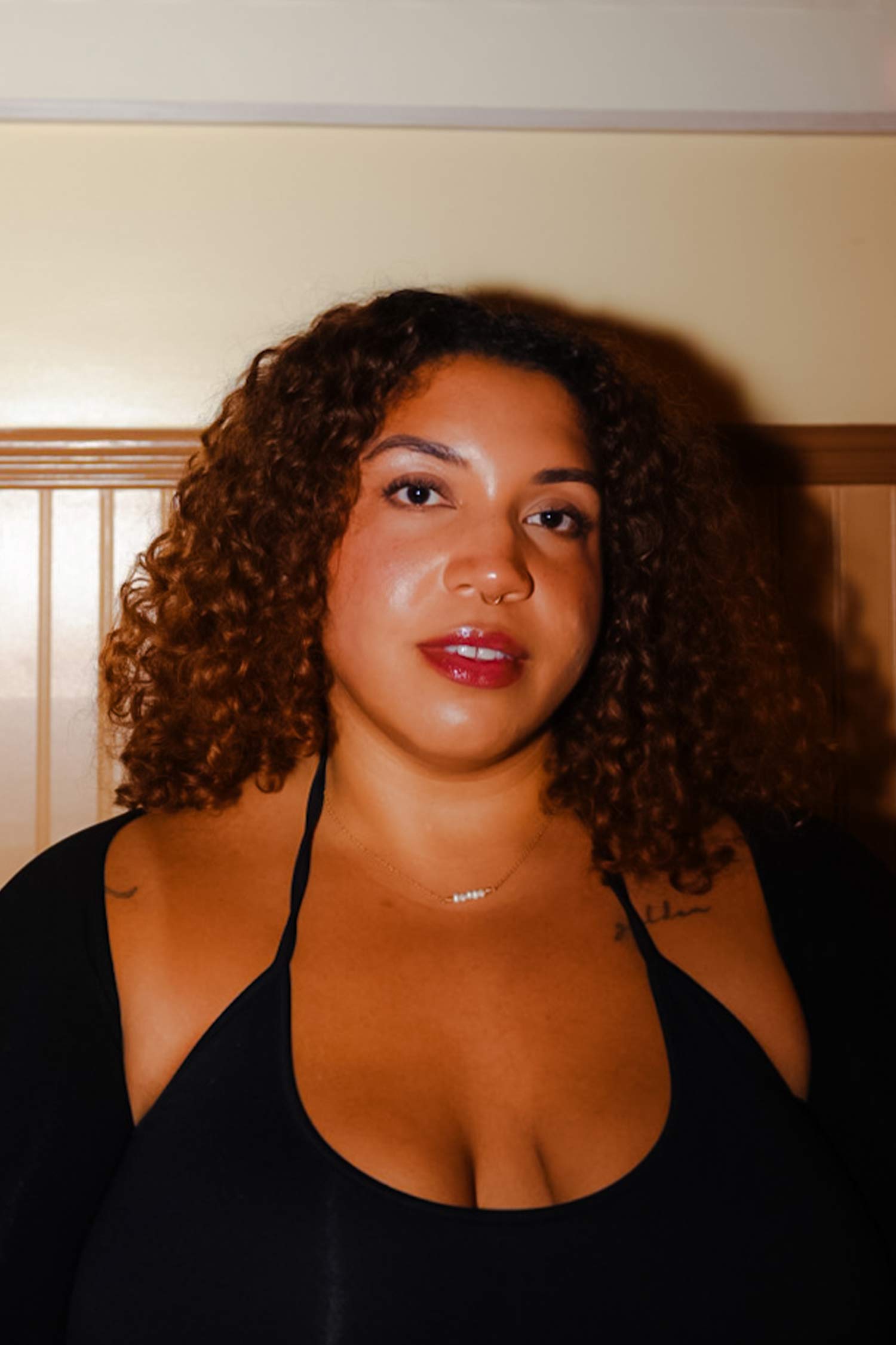 Portrait of a person with shoulder-length curly hair wearing a black top, photographed indoors against a wood-paneled wall.