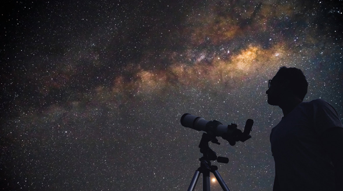 Silhouette of a person beside a telescope beneath a star-filled night sky and the Milky Way.