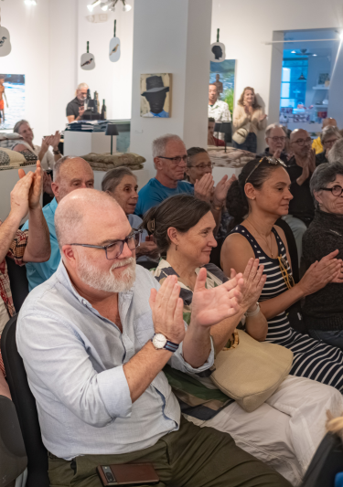 An audience of about two dozen adults seated in folding chairs, applauding at an event held in a bright gallery space displaying folded textiles and framed artwork.
