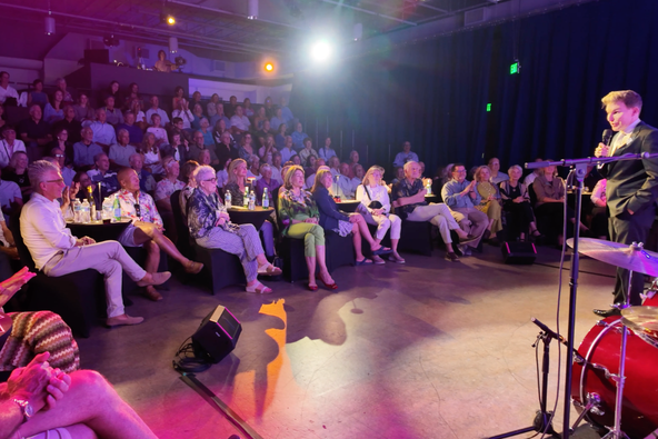 A performer in a dark suit sings into a microphone on a small stage as a seated audience watches from cabaret tables under pink and purple light.