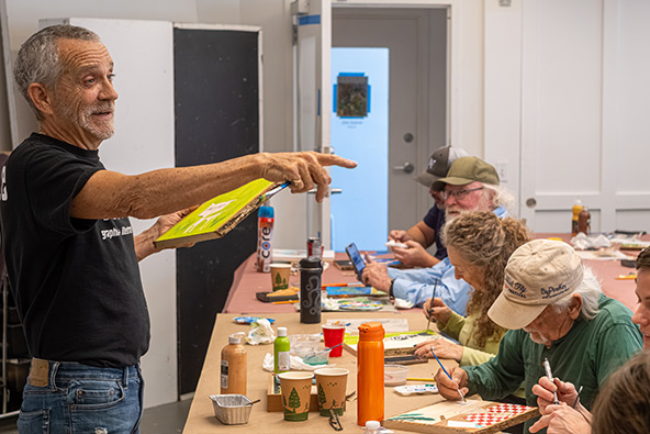 Instructor Andy Thurber gestures while teaching a group of adults painting and woodcarving together around a classroom table.