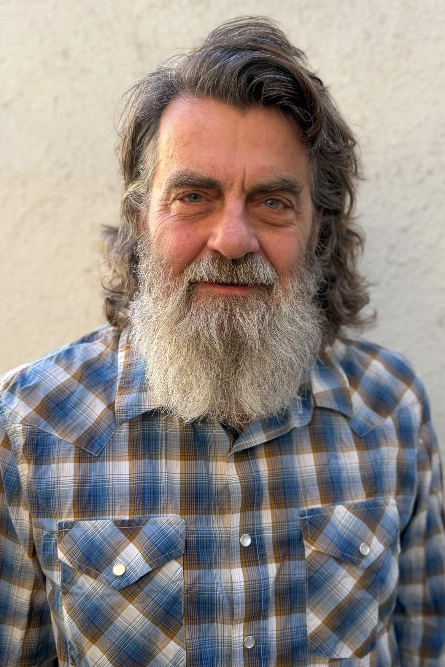 Portrait of a man with wavy gray hair and a long gray beard, wearing a blue, white and brown plaid shirt and smiling gently against a light-colored wall.