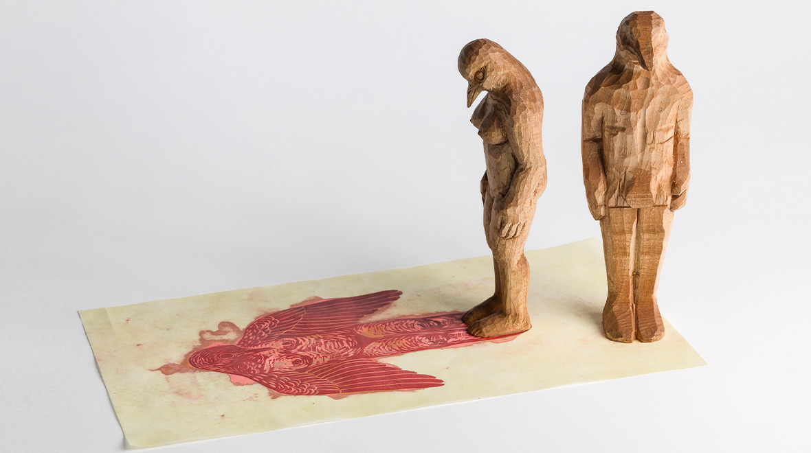 Wood sculpture of two standing human figures, with a bird-like head, positioned over a red printed image of a fallen winged figure.
