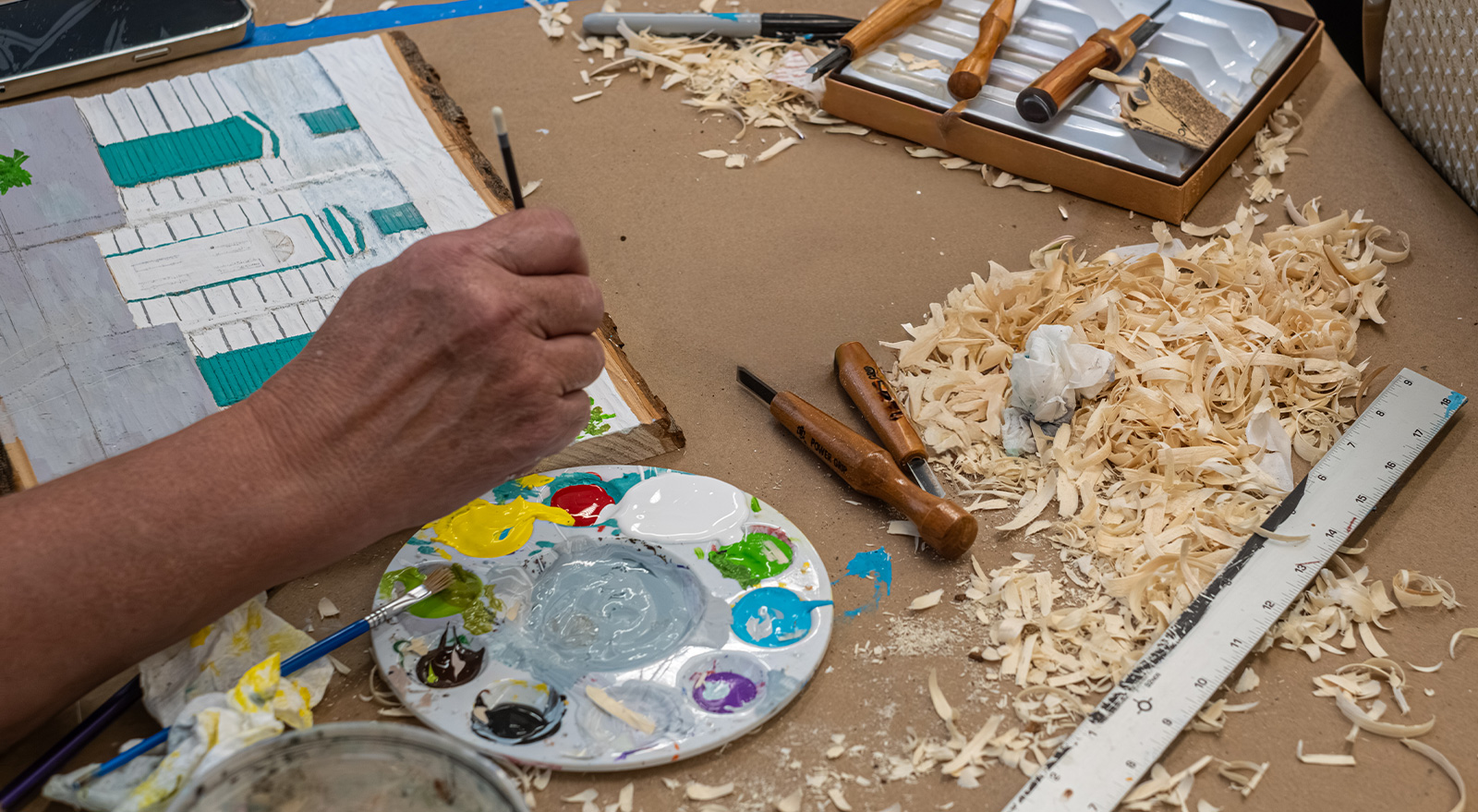 An artist paints a detailed architectural scene at a worktable surrounded by woodcarving tools, paint, rulers, and fresh wood shavings.