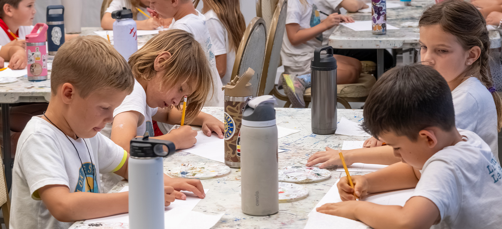 Children concentrate on drawing projects together around paint-splattered classroom tables during a youth art program.
