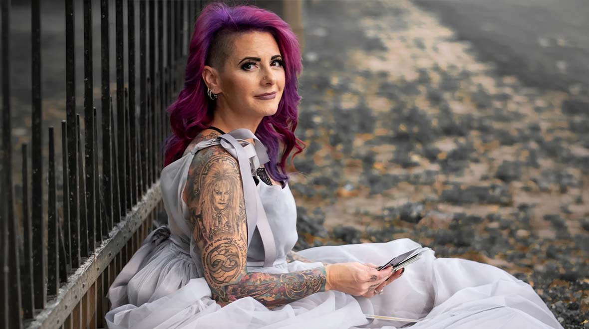 Woman with vivid purple hair and tattooed arms seated outdoors in a flowing gray dress, holding a deck of tarot cards.