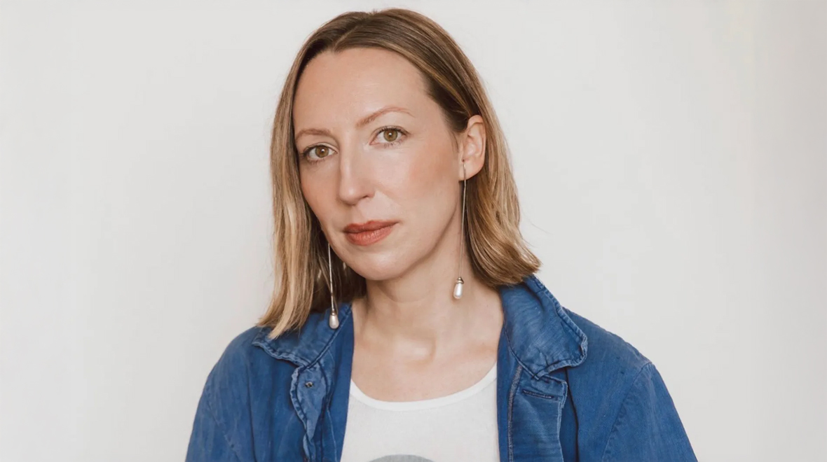 Portrait of a woman with shoulder-length light brown hair wearing a blue denim jacket and long pearl earrings against a white background.