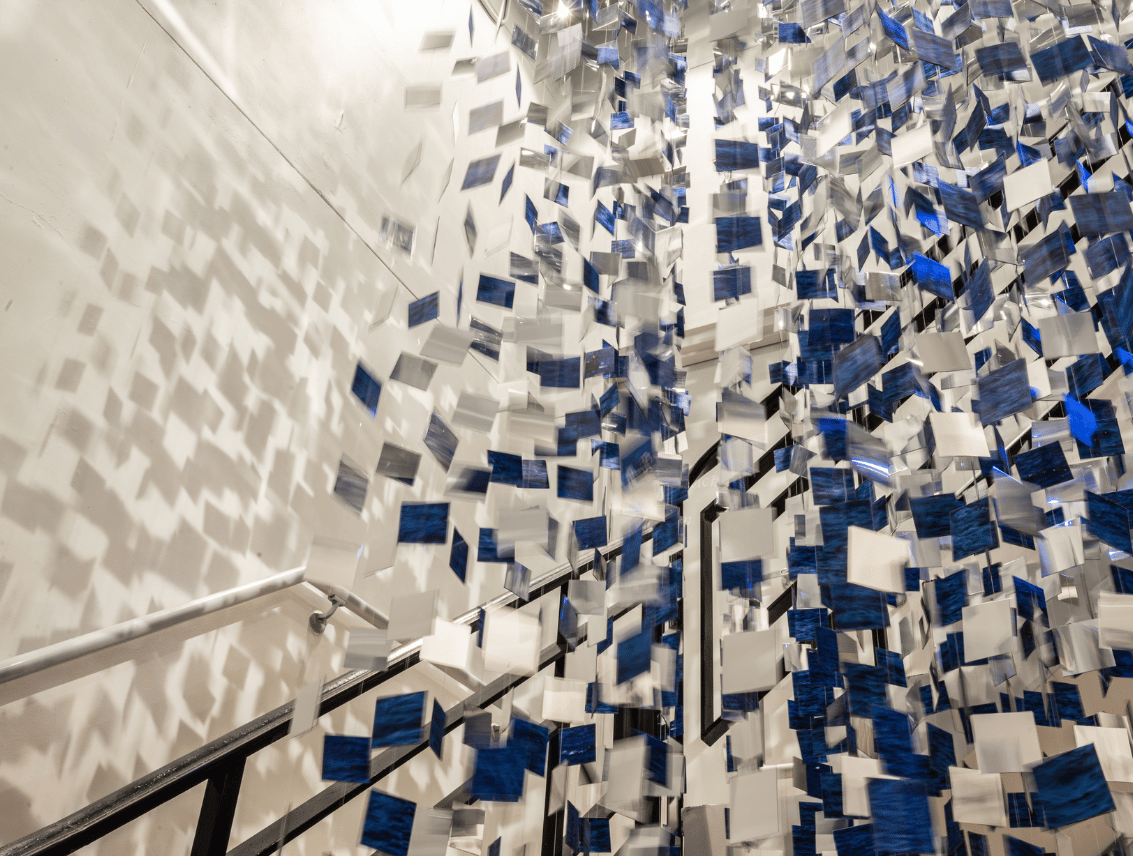 Immersive art installation filling a stairwell with hundreds of suspended blue, white, and translucent rectangular pieces, casting layered geometric shadows across the walls and stairs.
