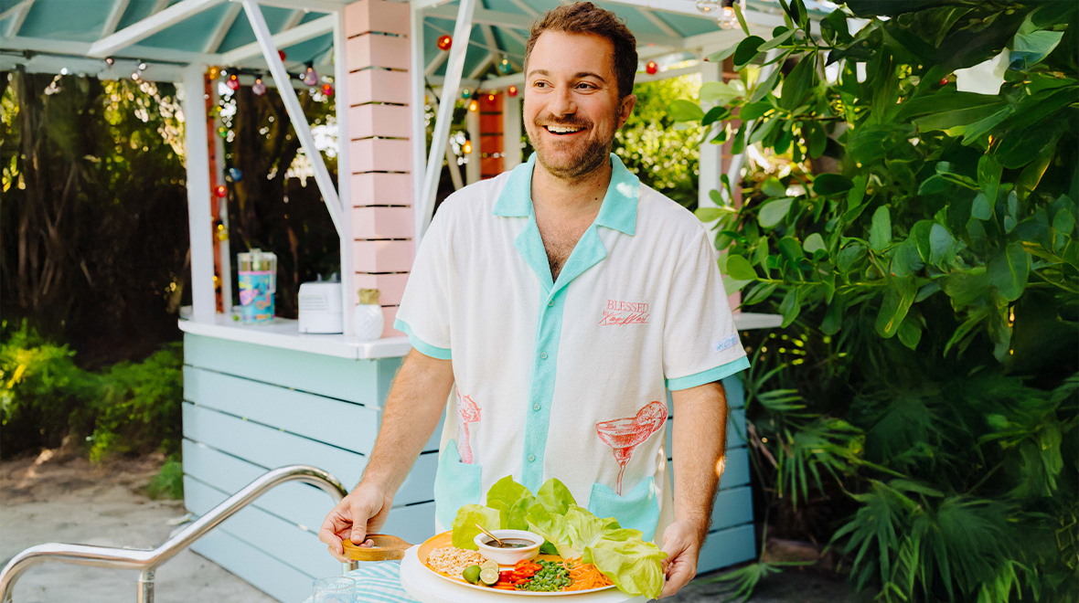 Smiling man holding a platter of food beside a pastel outdoor bar surrounded by tropical greenery.