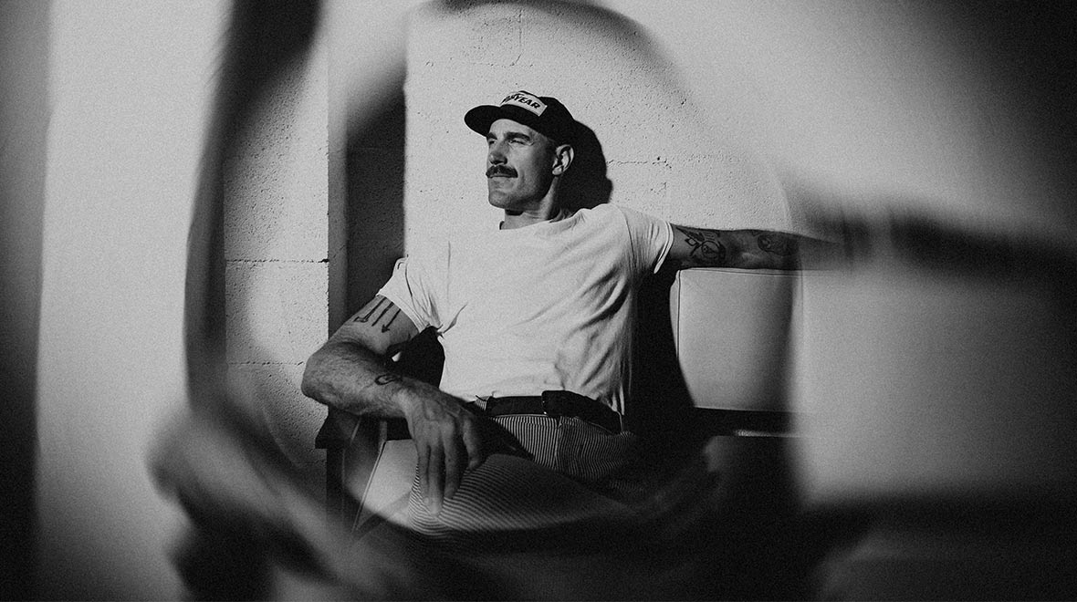 Black-and-white portrait of musician Rayland Baxter wearing a baseball cap and white T-shirt, seated with his tattooed arms outstretched against a wall, framed by soft shadows.